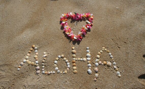 Destination Hawaii - Aloha written in the sand with shells and a heart shaped flower lei