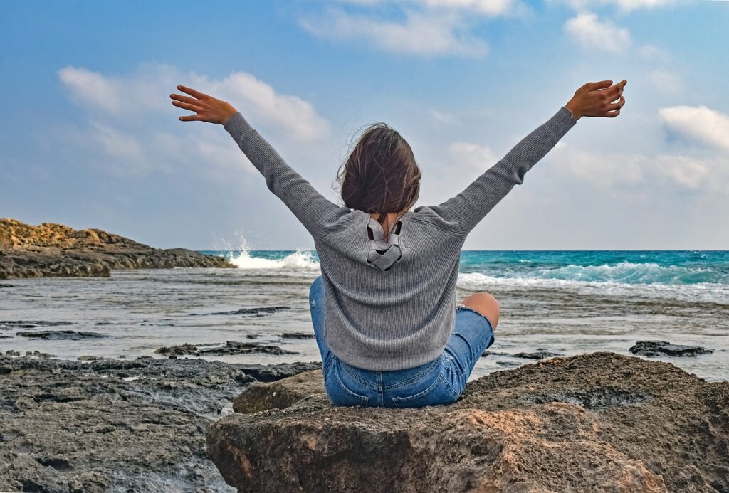 Wellness travel - woman with arms in the air looking out over the beach at the ocean