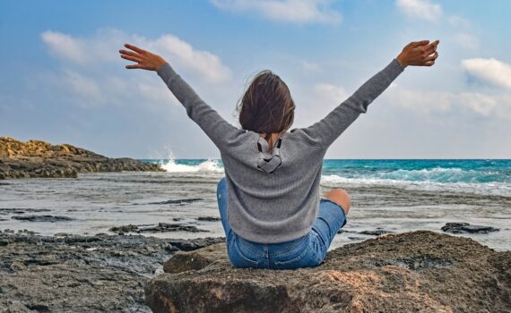 Wellness travel - woman with arms in the air looking out over the beach at the ocean