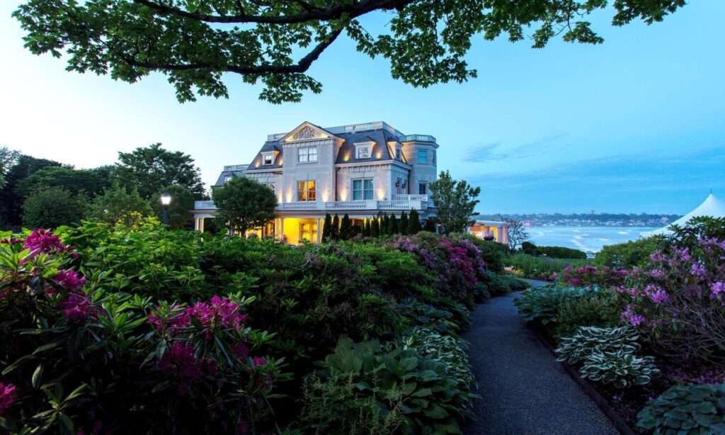 The Chanler at Cliff Walk - sun setting, hotel is lite up, ocean view behind the hotel, blooming rhododendrons in front of the hotel - a luxury hotel in Newport