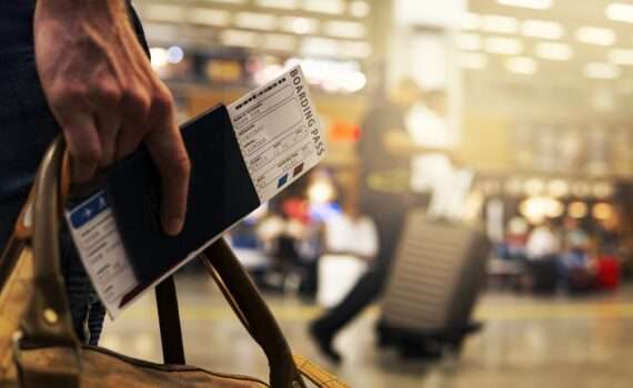 A travelers hand, in the airport holding a passport and a plane ticket and a carryon bag